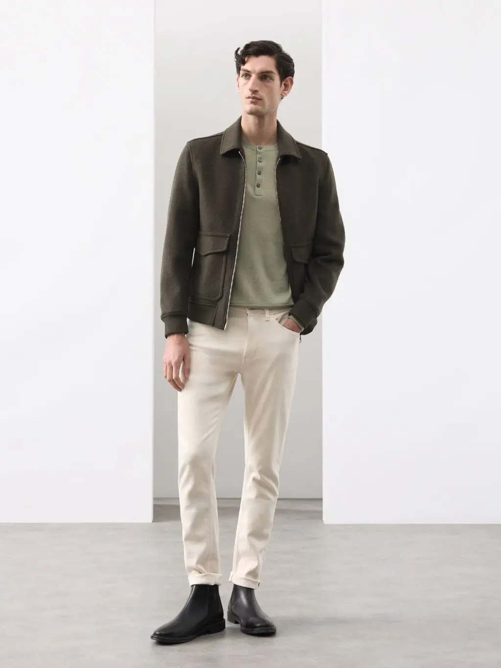 A man wears a pale green jumper, cream jeans and a leather jacket and boots