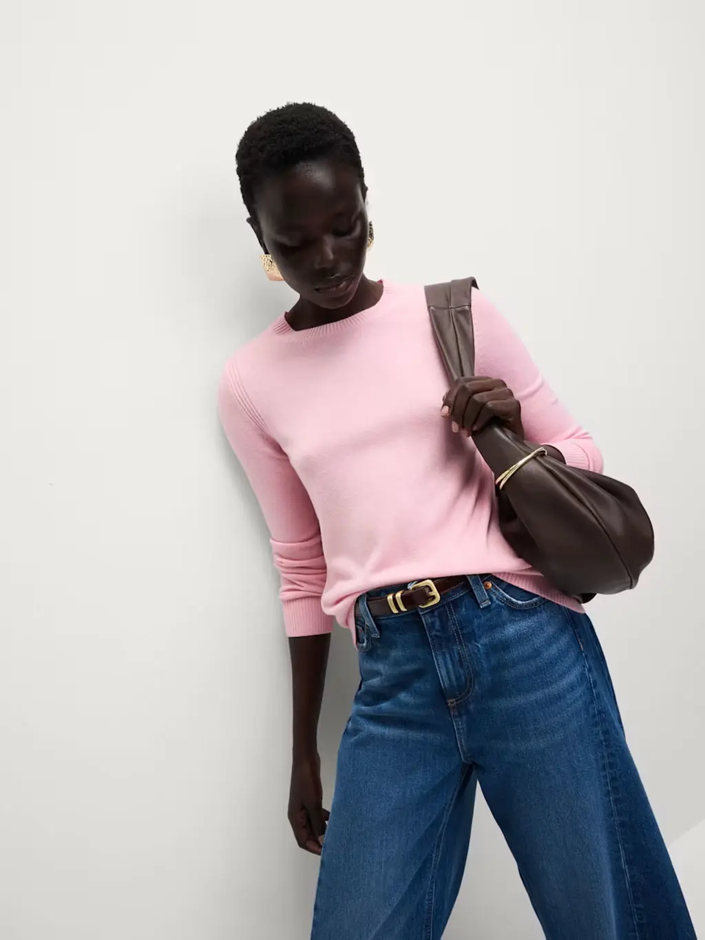 Woman wearing a pink jumper with blue trousers, a brown belt, brown bag and gold jewellery