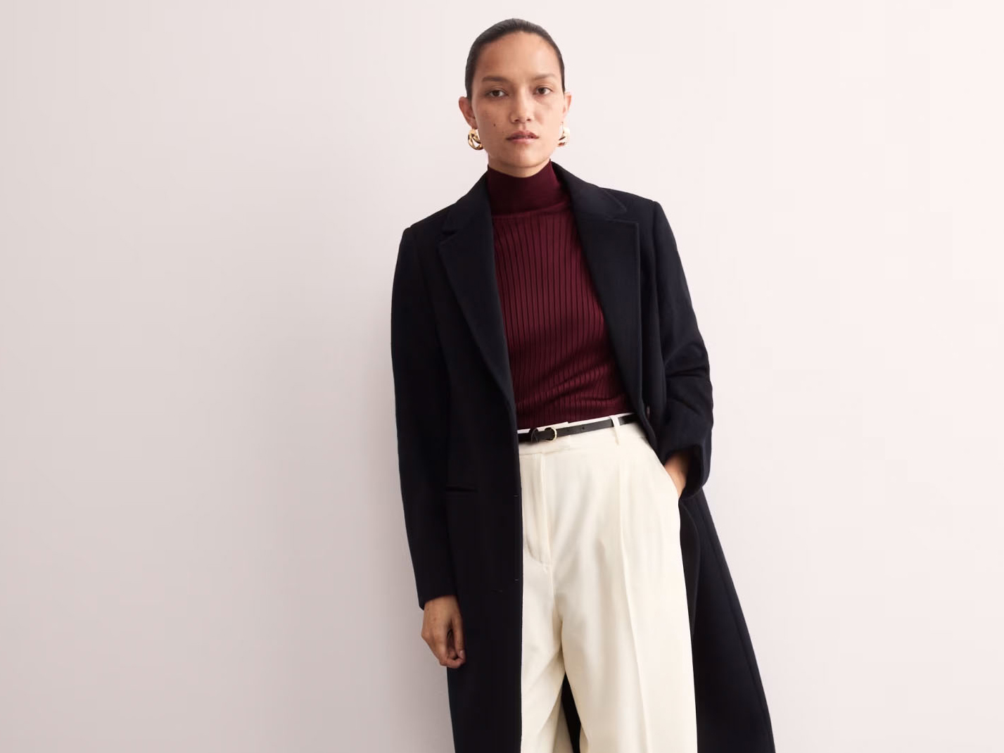 Woman wearing a 'quiet luxury' outfit, featuring a black Jaeger coat, burgundy top and cream tailored trousers