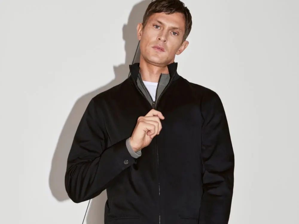 A man zipping up a black bomber jacket while stood against a white wall