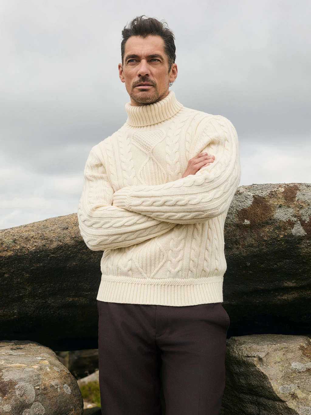 Man wearing a cream cable knit jumper and brown trousers leaning against a rock.