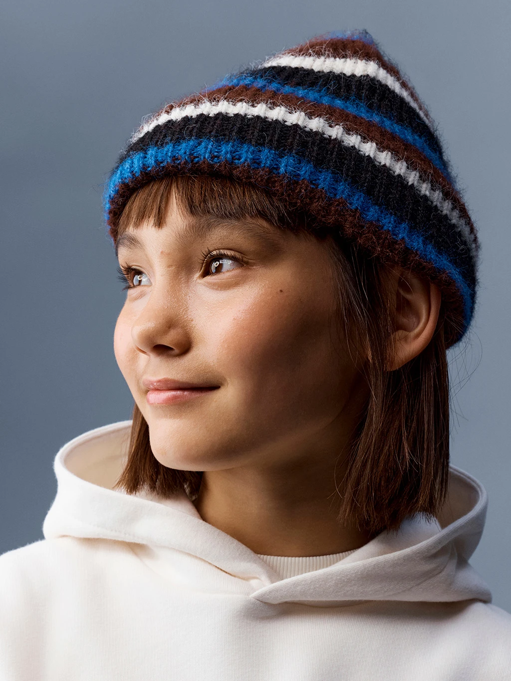 Young child in a cream hoodie and blue, white and red stripy knitted hat.