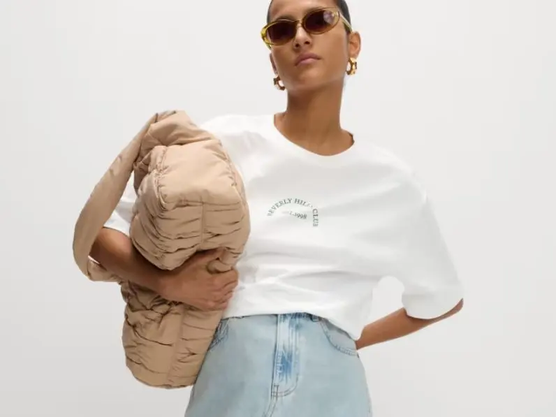 A woman wears peach sunglasses, an oversized white T-shirt and holds a quilted handbag under her arm