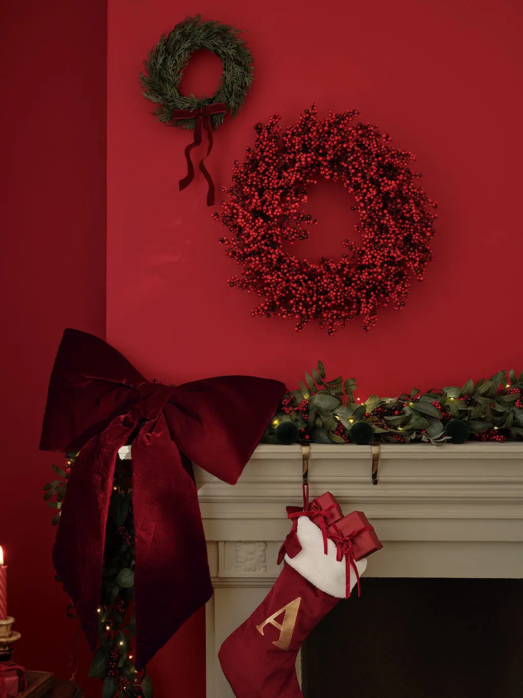 A red wall and mantlepiece decorated with one green and one red Christmas wreath, alongside a bow and stocking