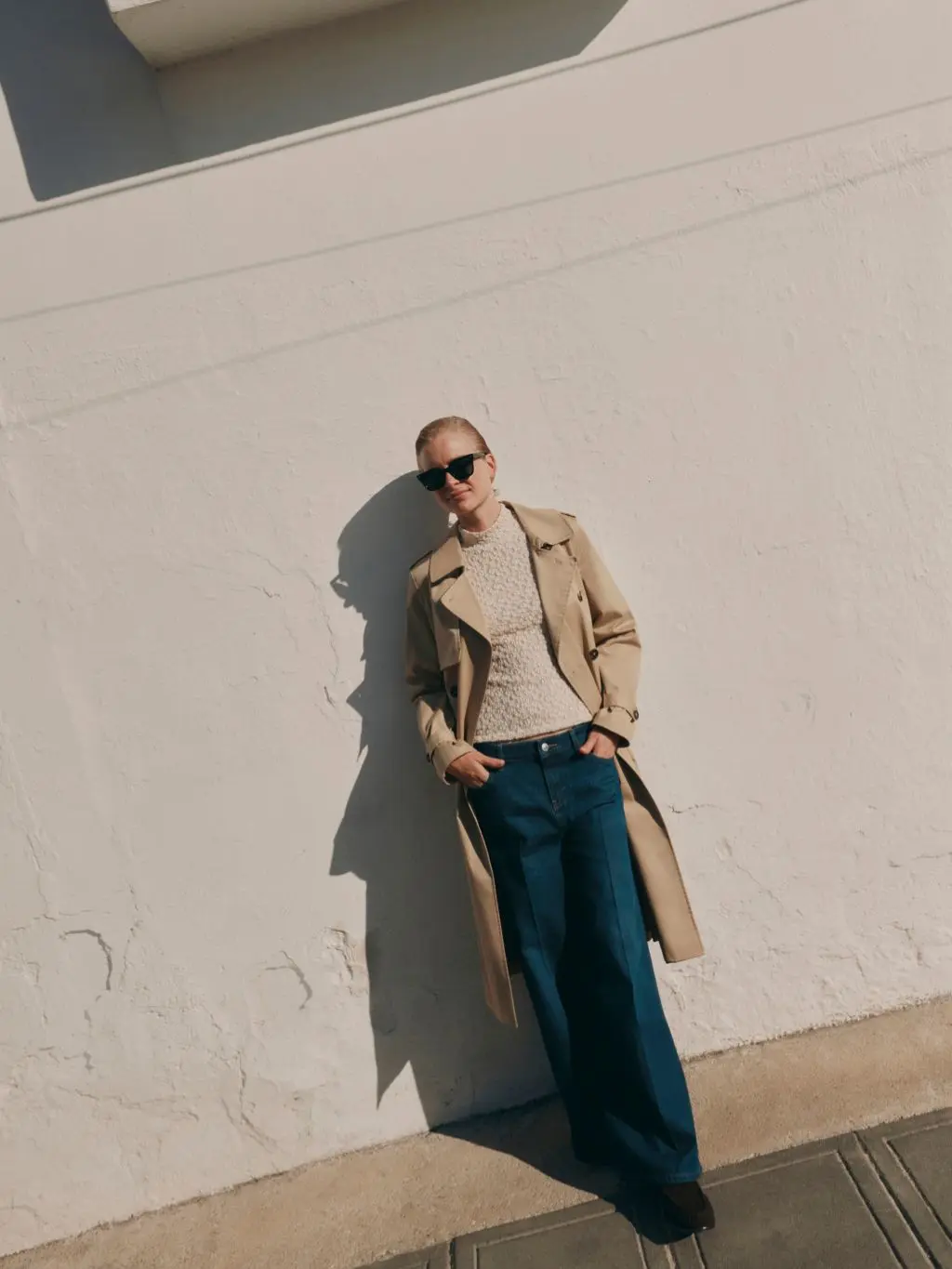 A model leans against a white wall wearing a beige trench coat, cream top and dark blue jeans
