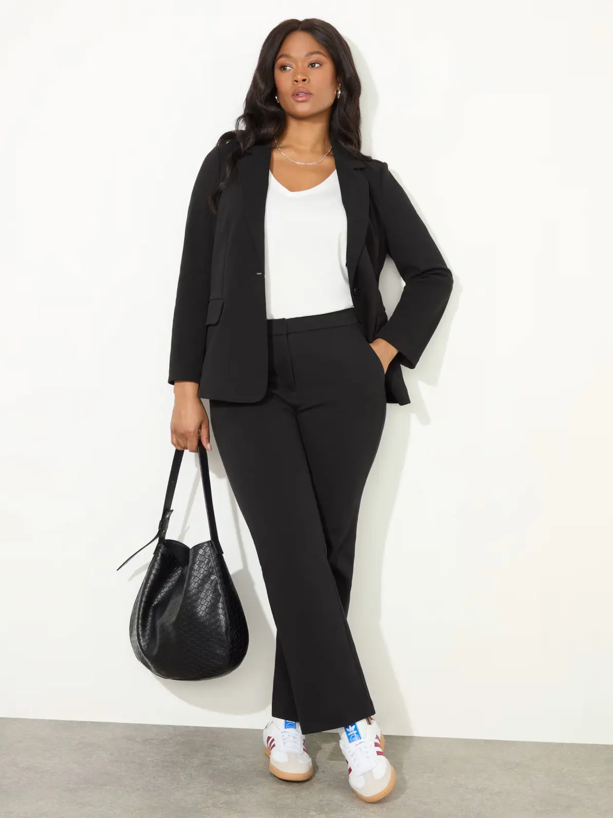 Woman wearing black blazer over white V-neck top, black smart trousers and white trainers, holding black bag