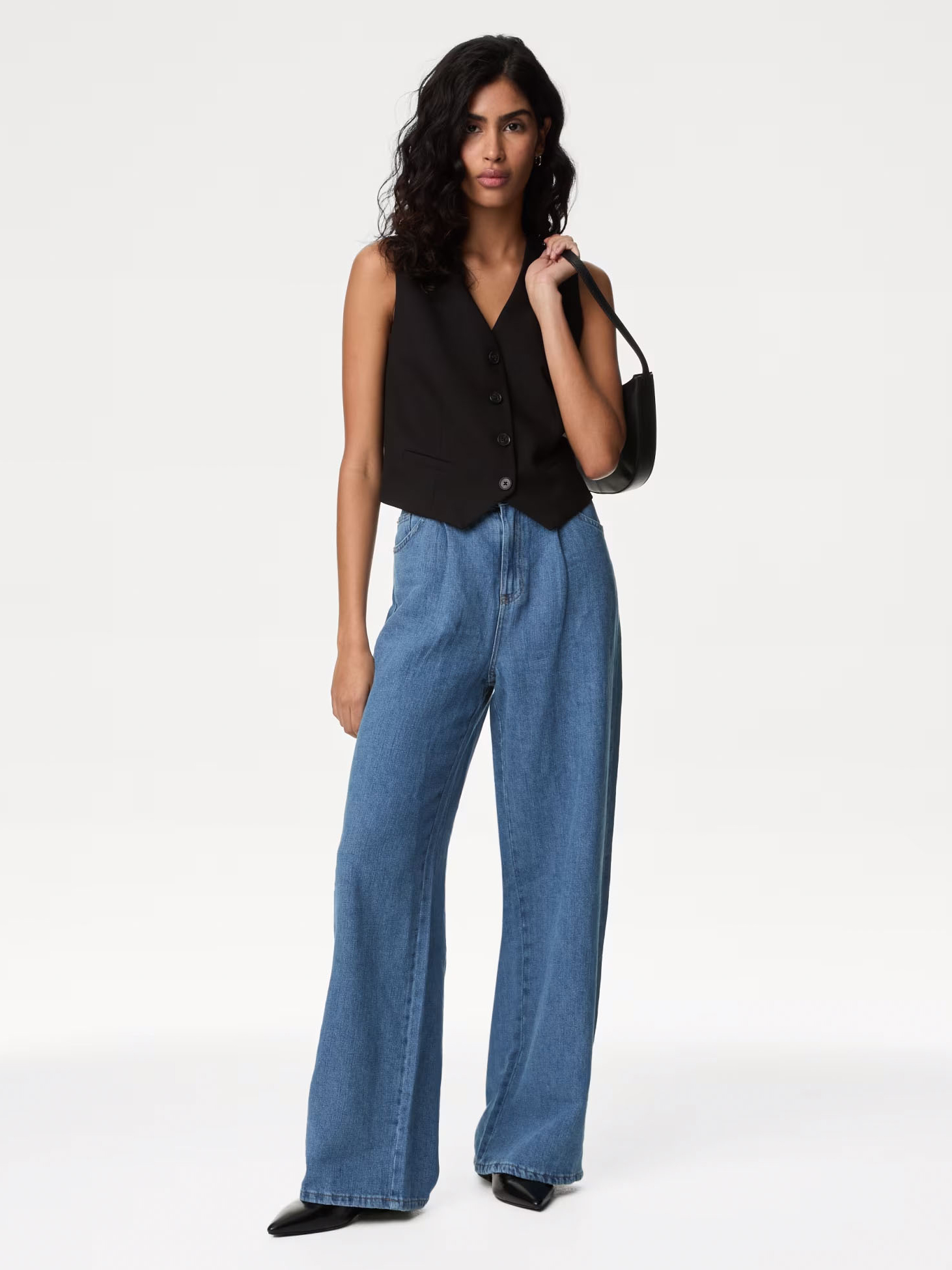 Woman wearing black waistcoat, blue wide-leg jeans and a black shoulder bag. Shop wide-leg jeans