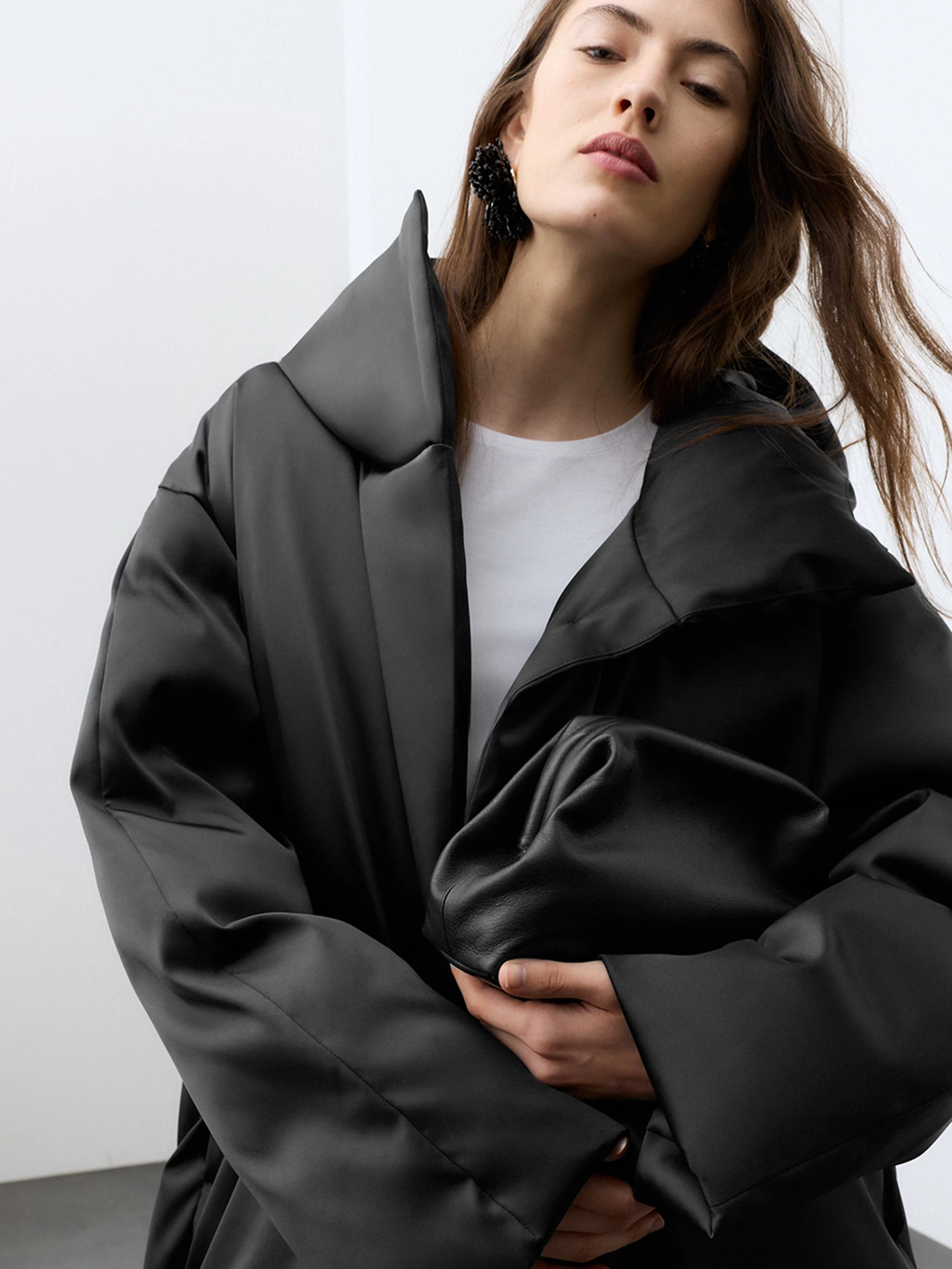 Woman wearing a black satin puffer coat with a white top underneath, holding a black clutch bag.