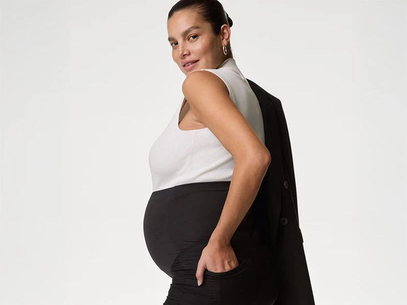 Pregnant woman wearing a white high-neck top with black leggings, a blazer and silver earrings.