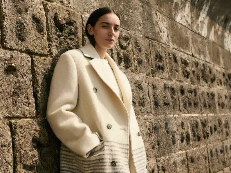 A woman wearing a cream wool coat by Jaeger leans against a sea wall