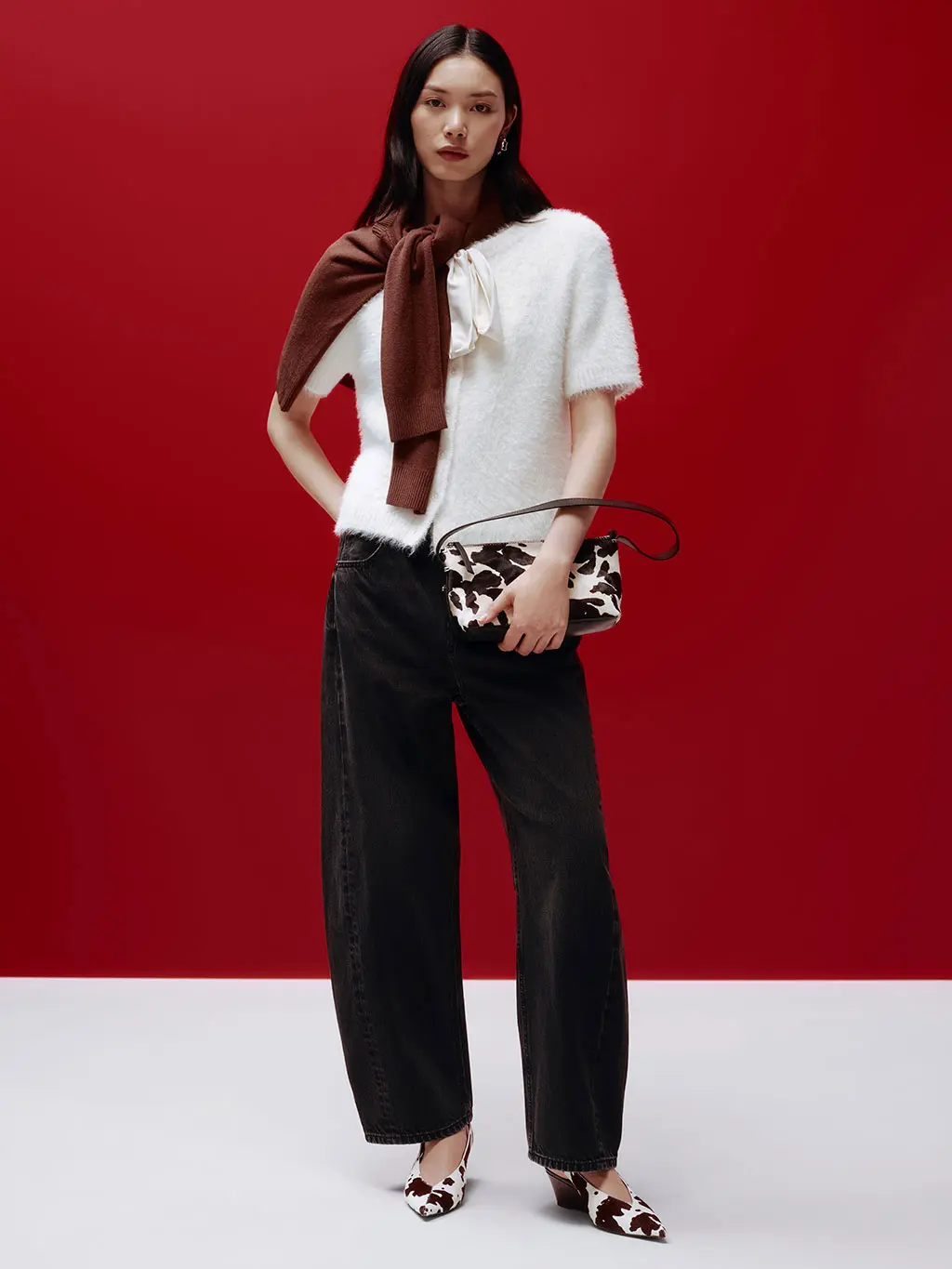 Woman wearing black barrel-leg jeans with a white cardigan, brown jumper over the shoulders and animal-print heels and bag