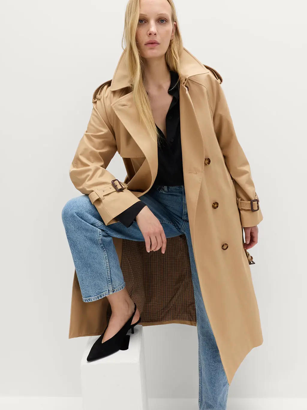 Woman wearing a trench coat over a black blouse, blue jeans and black kitten heels