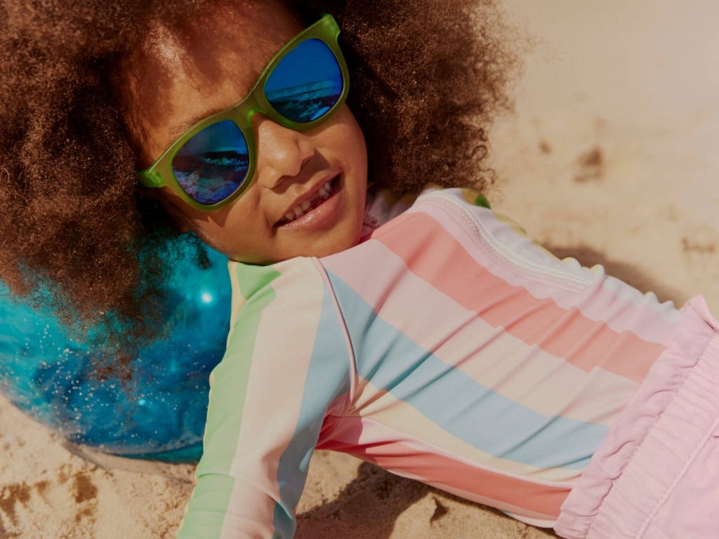 Girl wearing a long-sleeved multi-coloured striped swimsuit with pink shorts and green sunglasses. Shop girls’ swimwear