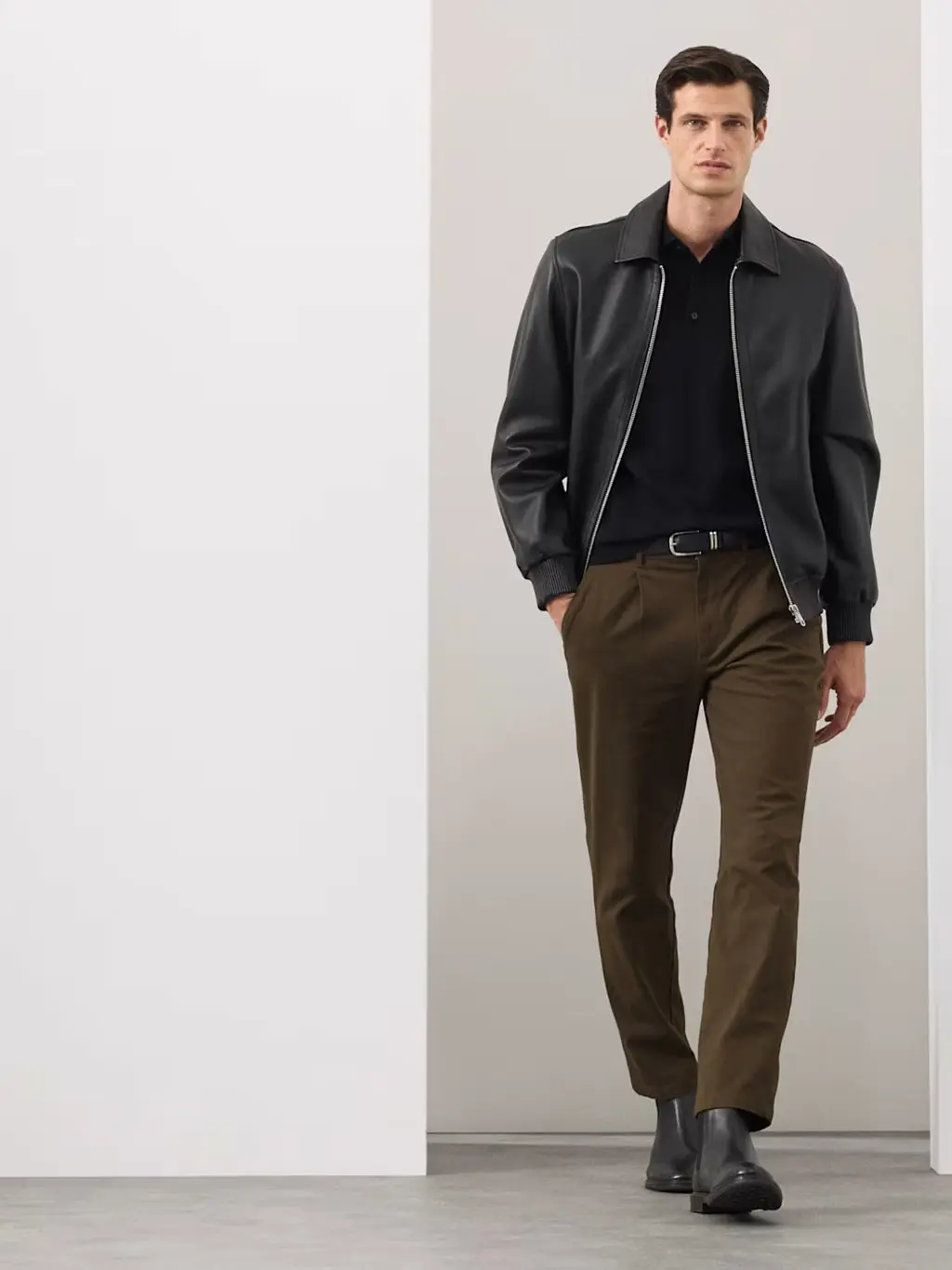 Man wearing khaki pleated chinos with a black polo shirt, black leather jacket, black belt and Chelsea boots