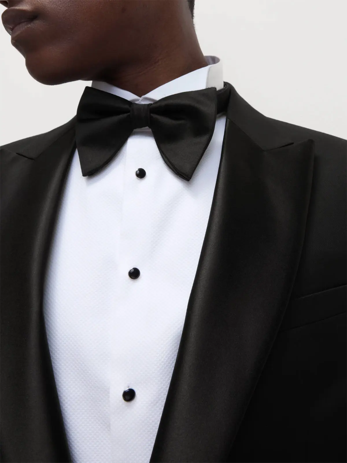 Man wearing black blazer over white shirt with black buttons, and black bow tie 