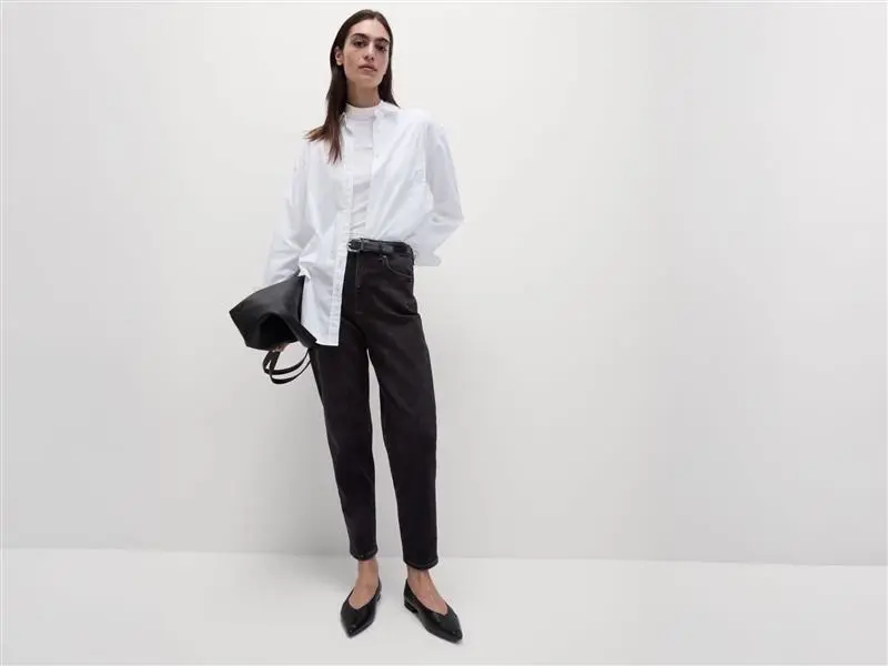 Woman wearing black mom jeans with a white T-shirt, white shirt, black ballet pumps and a black bag.