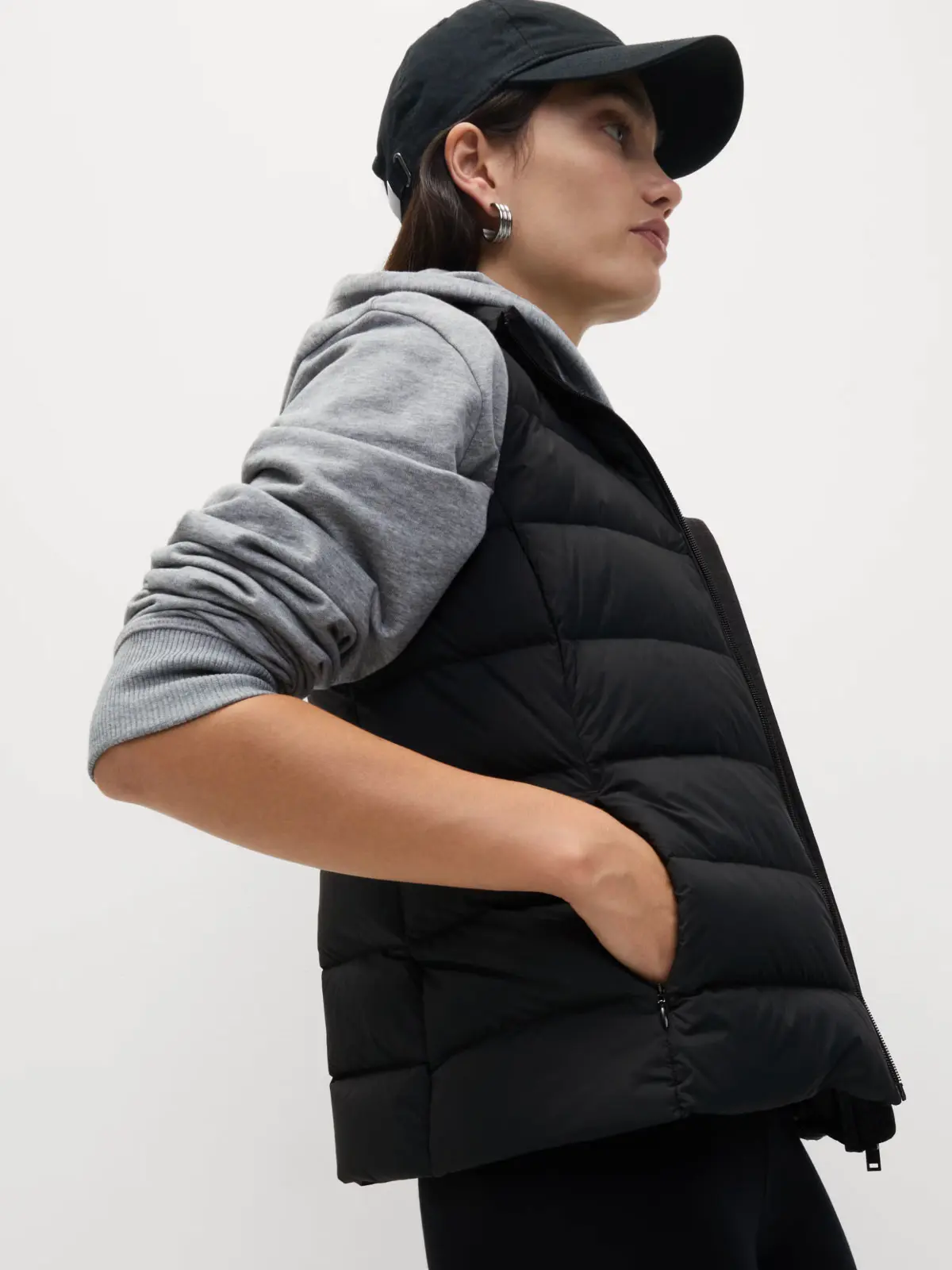 Woman wearing black gilet over grey sweatshirt, styled with a black cap 