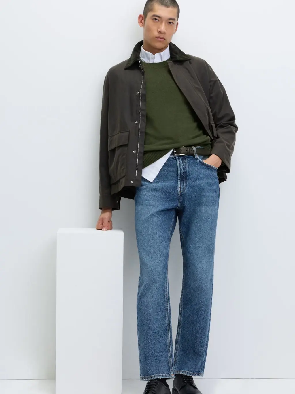 A man wears a green jumper over a shirt with jeans and a wax jacket