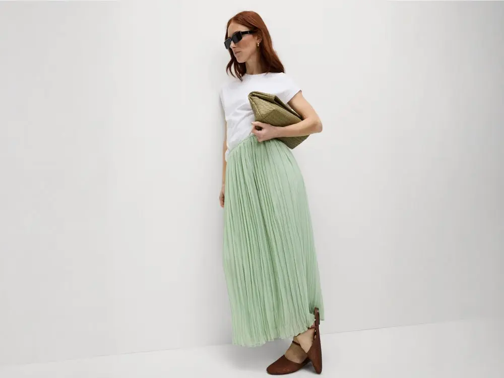 A woman leans against a wall wearing a white t-shirt, pistachio cream skirt, carrying an olive green clutch bag and wearing sunglasses
