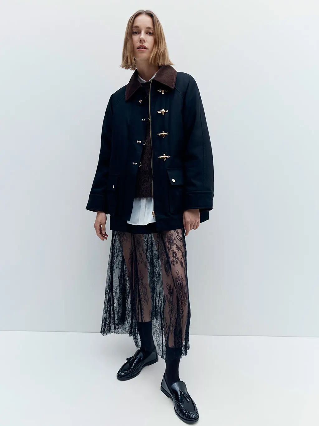 Woman wearing a black lace skirt with a white shirt, cardigan, barn jacket and loafers with socks