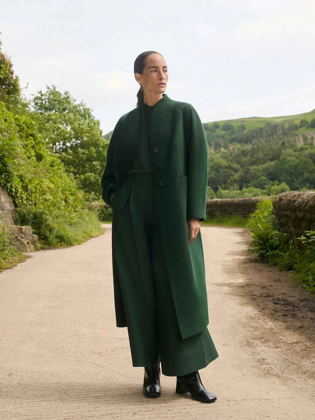 Woman wearing a dark green wool long coat stood on a path in the countryside.