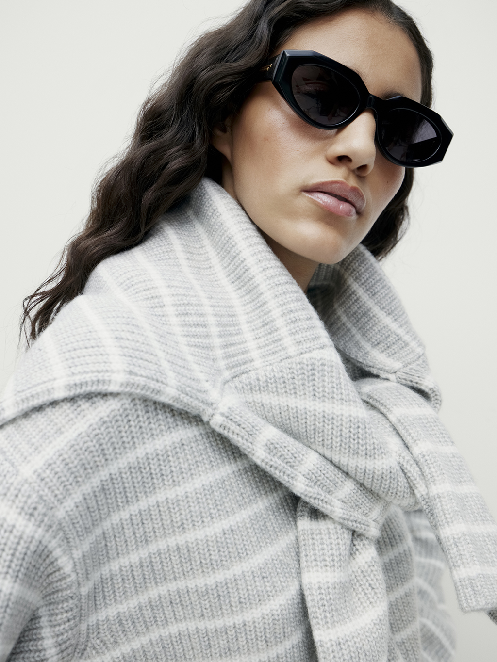 Woman wearing grey striped jumper with a grey striped jumper knotted around her shoulders and black sunglasses