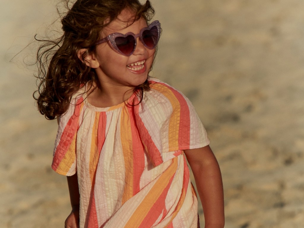 Girl wearing a short-sleeved cream, orange and pink striped dress with heart-shaped sunglasses. Visit The Holiday Shop
