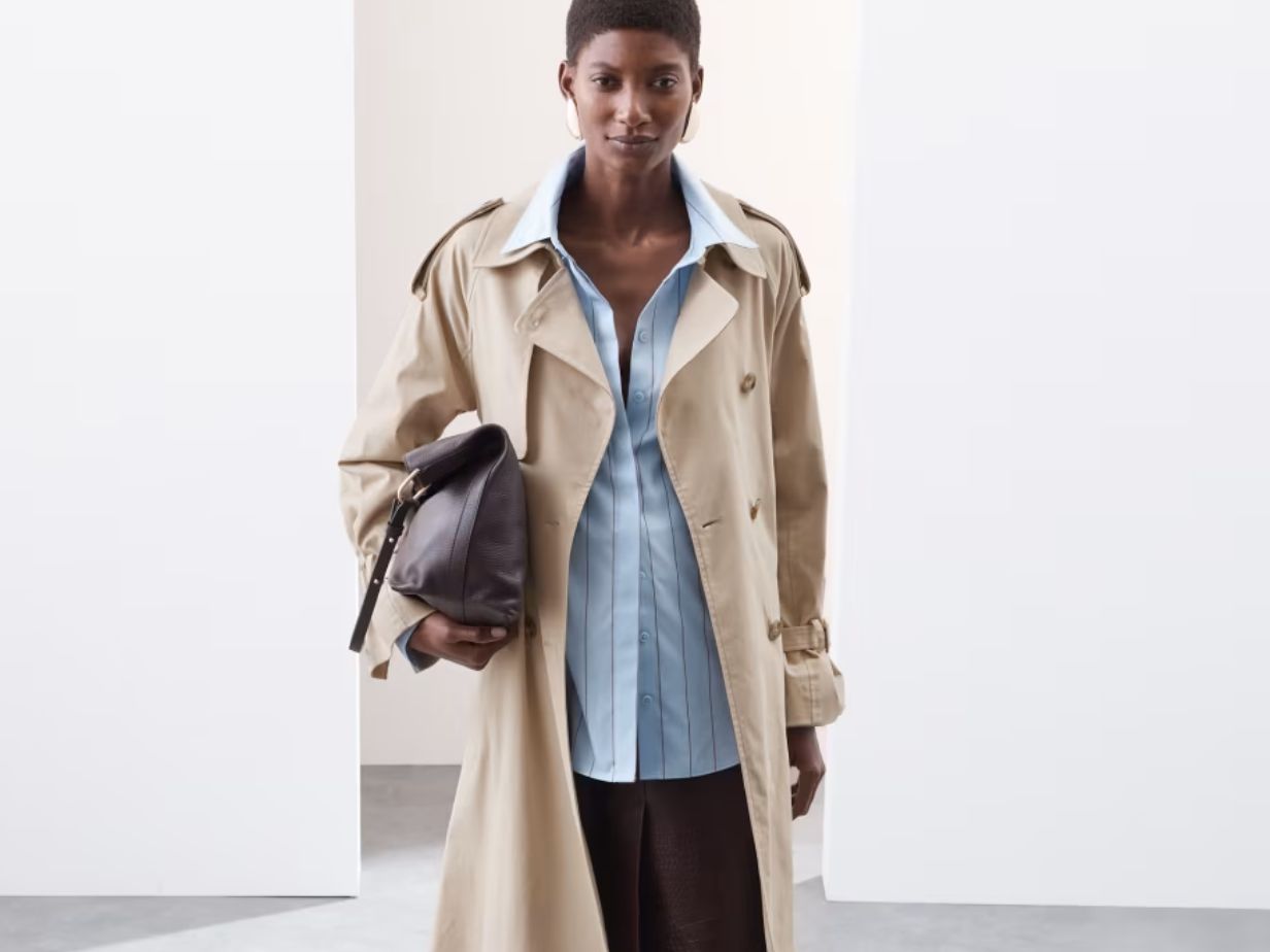 A woman wearing a beige trench over a light blue striped shirt, carrying a brown bag under her arm