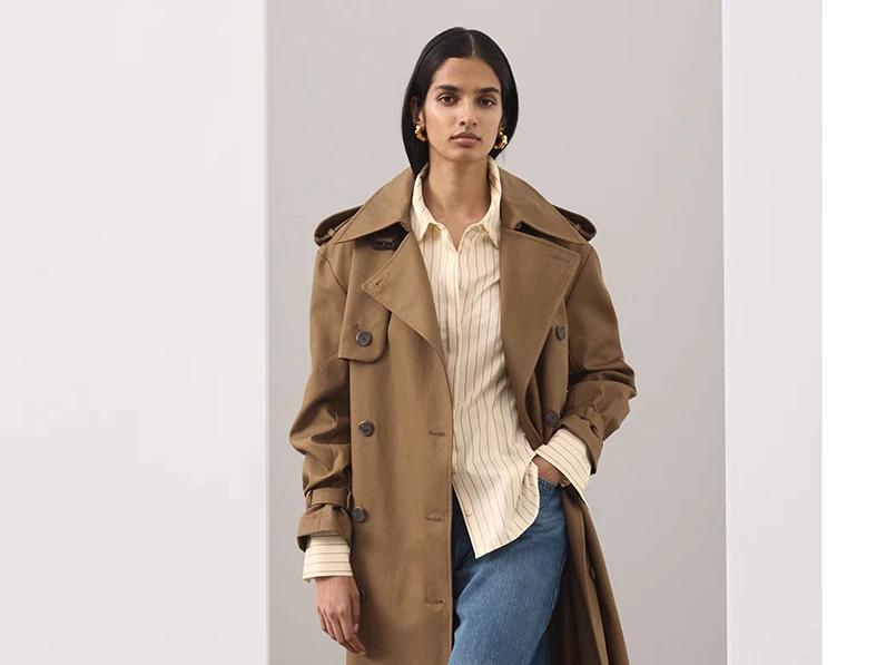 A woman wearing a beige trench, a light yellow striped shirt and blue jeans