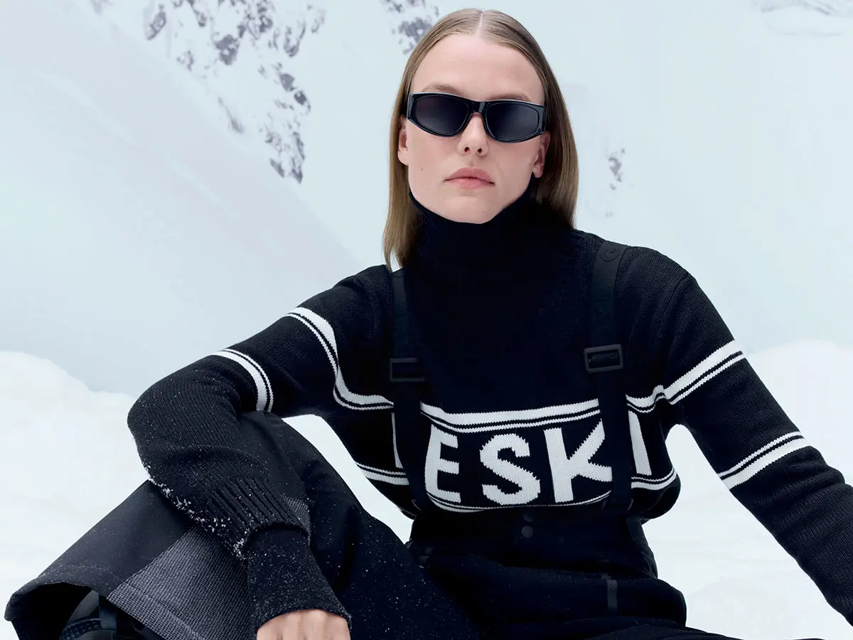 Woman sitting down wearing black jumper with white slogan, black ski trousers and ski glasses 