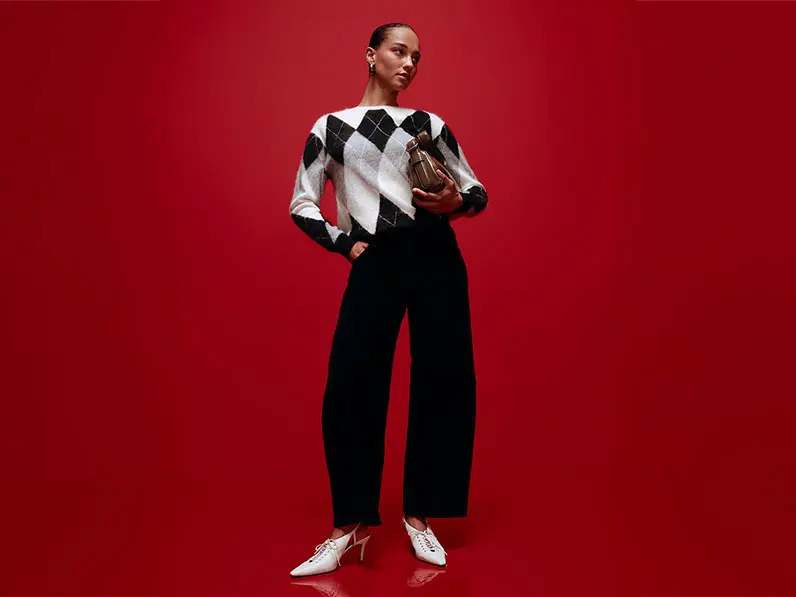 Woman wearing black barrel-leg jeans with an argyle jumper, white heels and a clutch bag