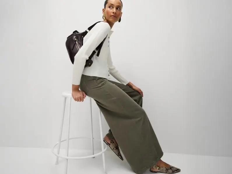 A woman is sat on a stool wearing a cream cardigan, khaki trousers and carrying a black handbag