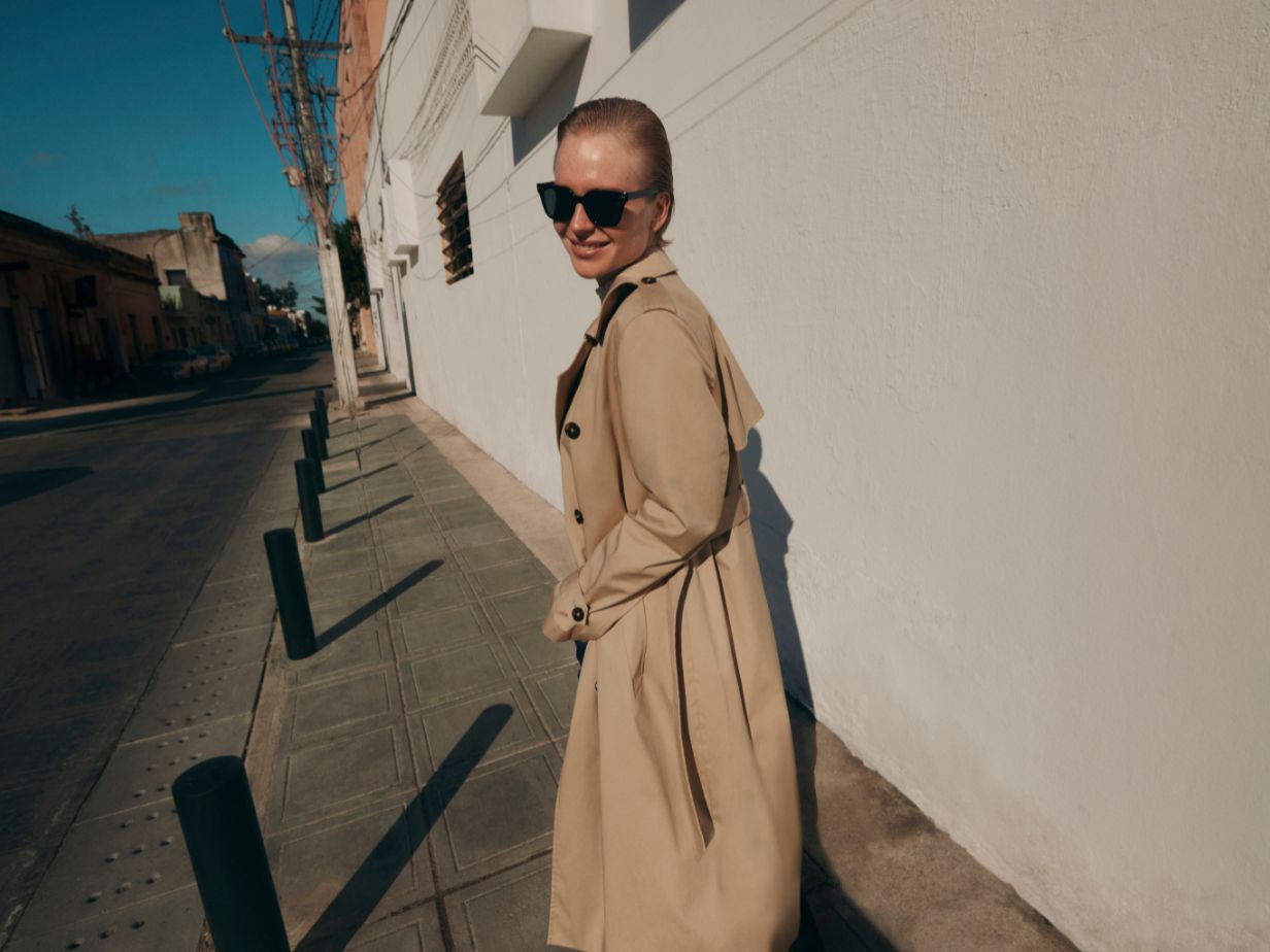 A model walks down a road in a trench coat and sunglasses on