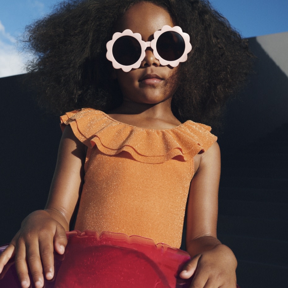 Girl wearing an orange frilled swimsuit and light pink floral sunglasses. Shop kids’ swimwear
