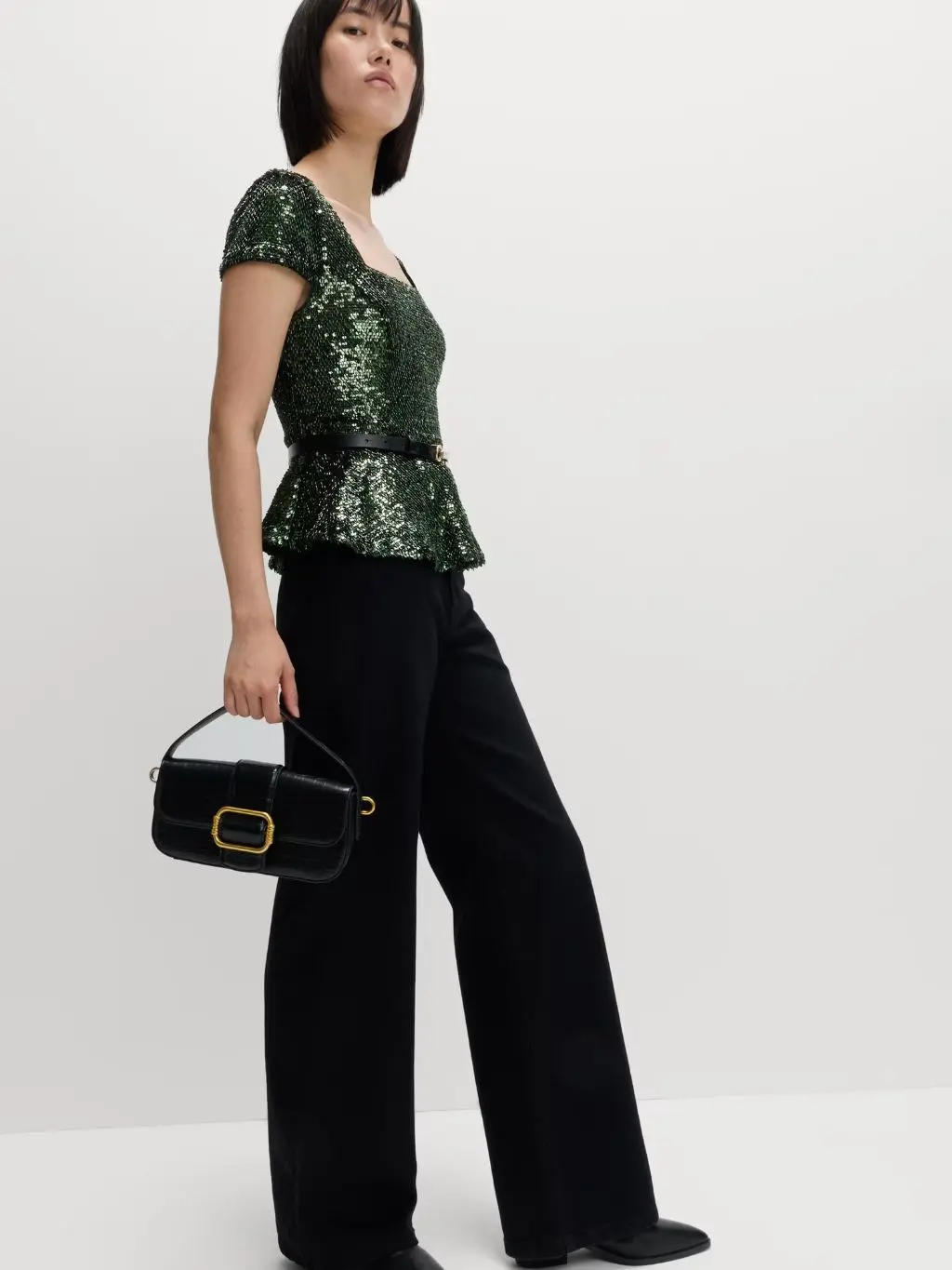 A woman wears a green sequin peplum top with black jeans and carries a baguette handbag