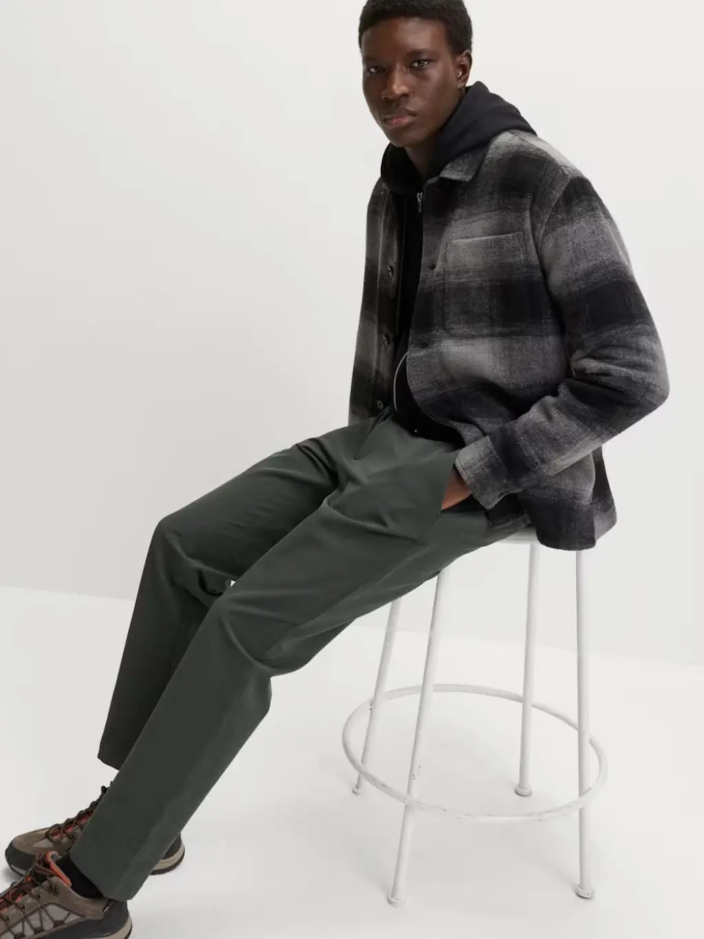 A man sits on a stool wearing dark grey chinos and a checked shirt over a hooded top