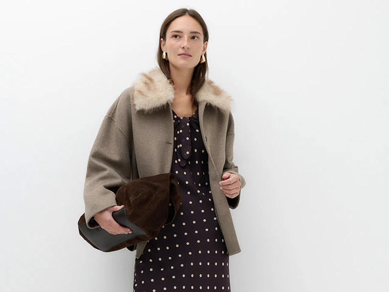Woman wearing a brown polka dot dress, beige coat with a fluffy collar, and suede shoulder bag.