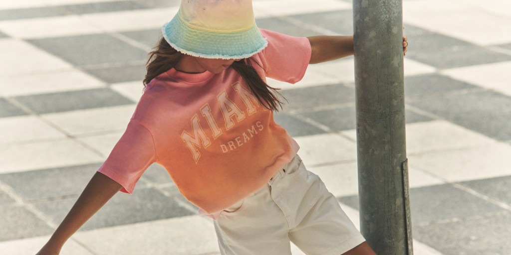 Girl wearing a bucket hat, pink graphic print T-shirt and beige shorts. Shop girls’ clothing