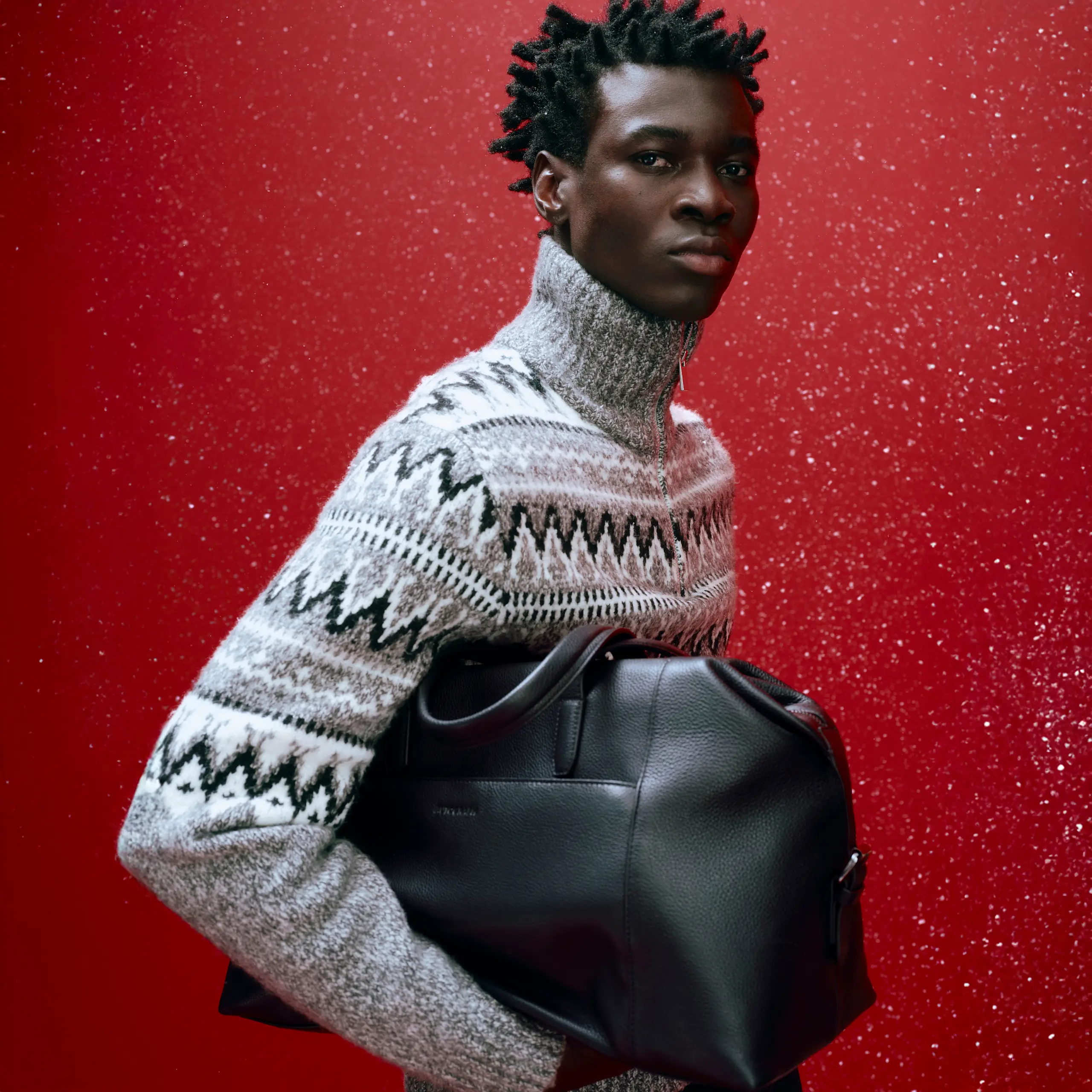 Man wearing a patterned quart-zip jumper with a black leather bag against a sparkly red backdrop
