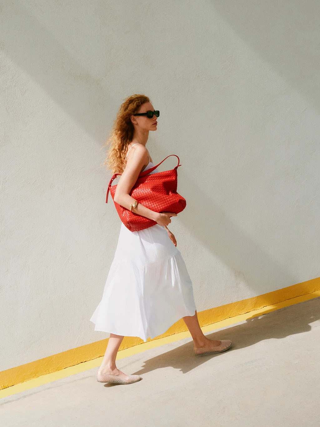 A woman wearing a white dress carries a woven red bag under one arm