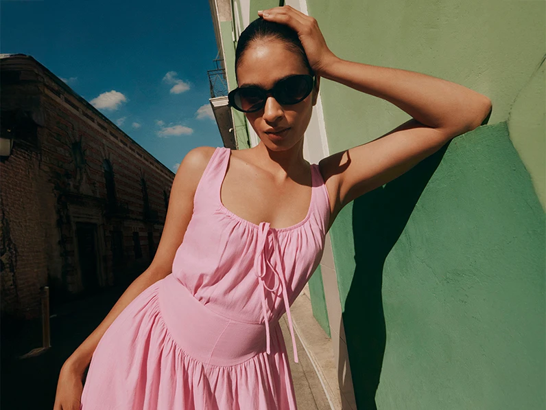 Woman wearing a pink linen-rich dress with black sunglasses by a green wall