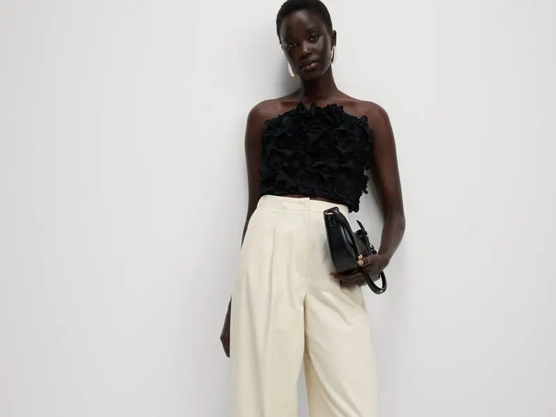 Woman wearing ecru barrel-leg trousers with a black strapless top, gold earrings and a black handbag.