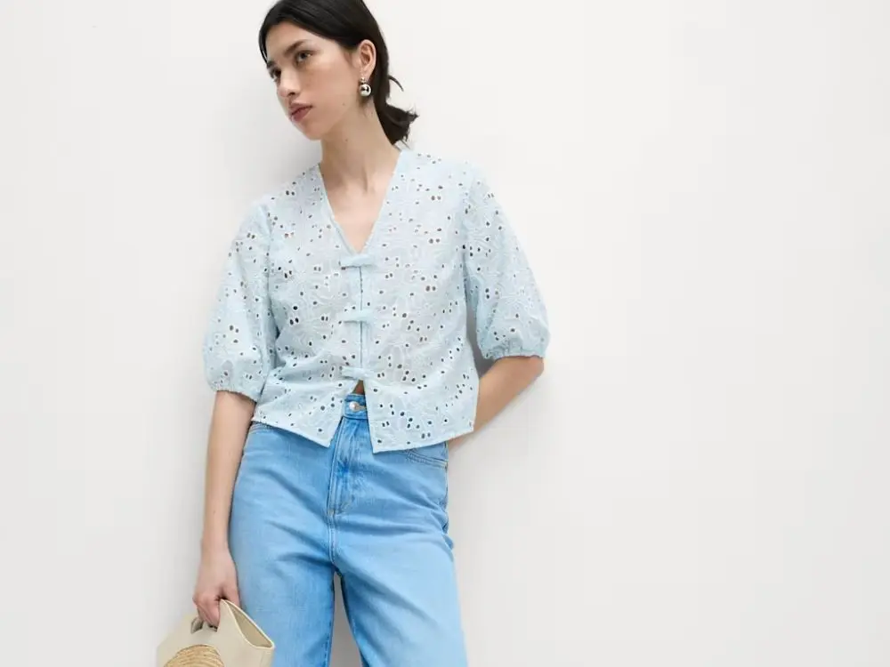 A woman wears a baby blue floral cutout blouse with jeans, carrying a straw bag