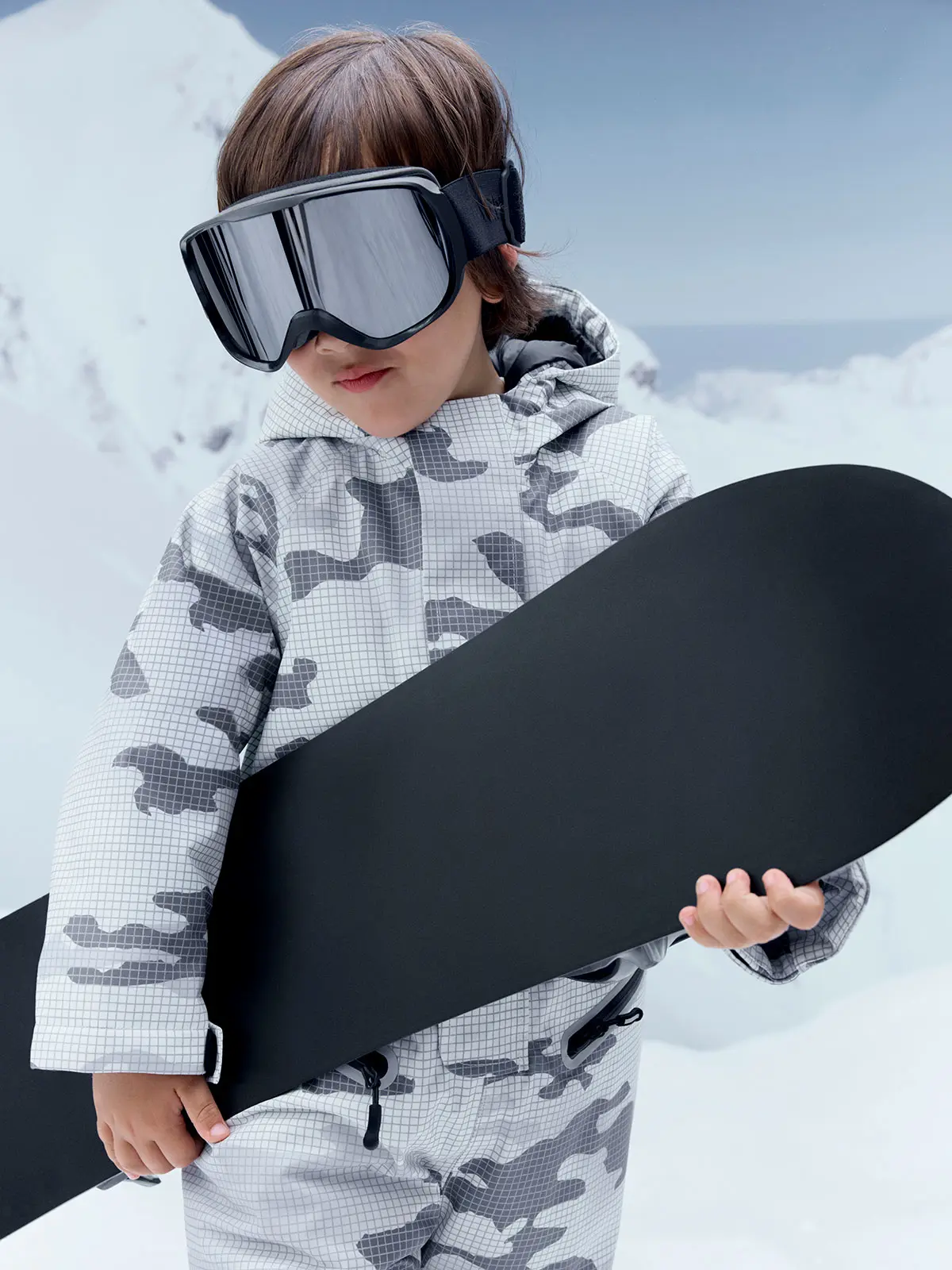 Boy wearing grey and white camouflage-print ski jacket, matching trousers and ski goggles. Holding black snowboard