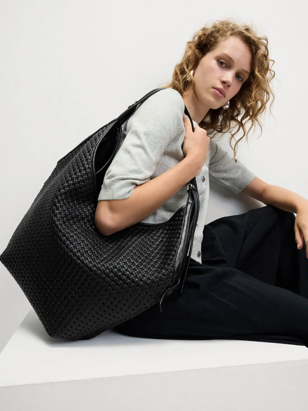 Woman wearing a grey jumper and black trousers with a black leather tote bag.