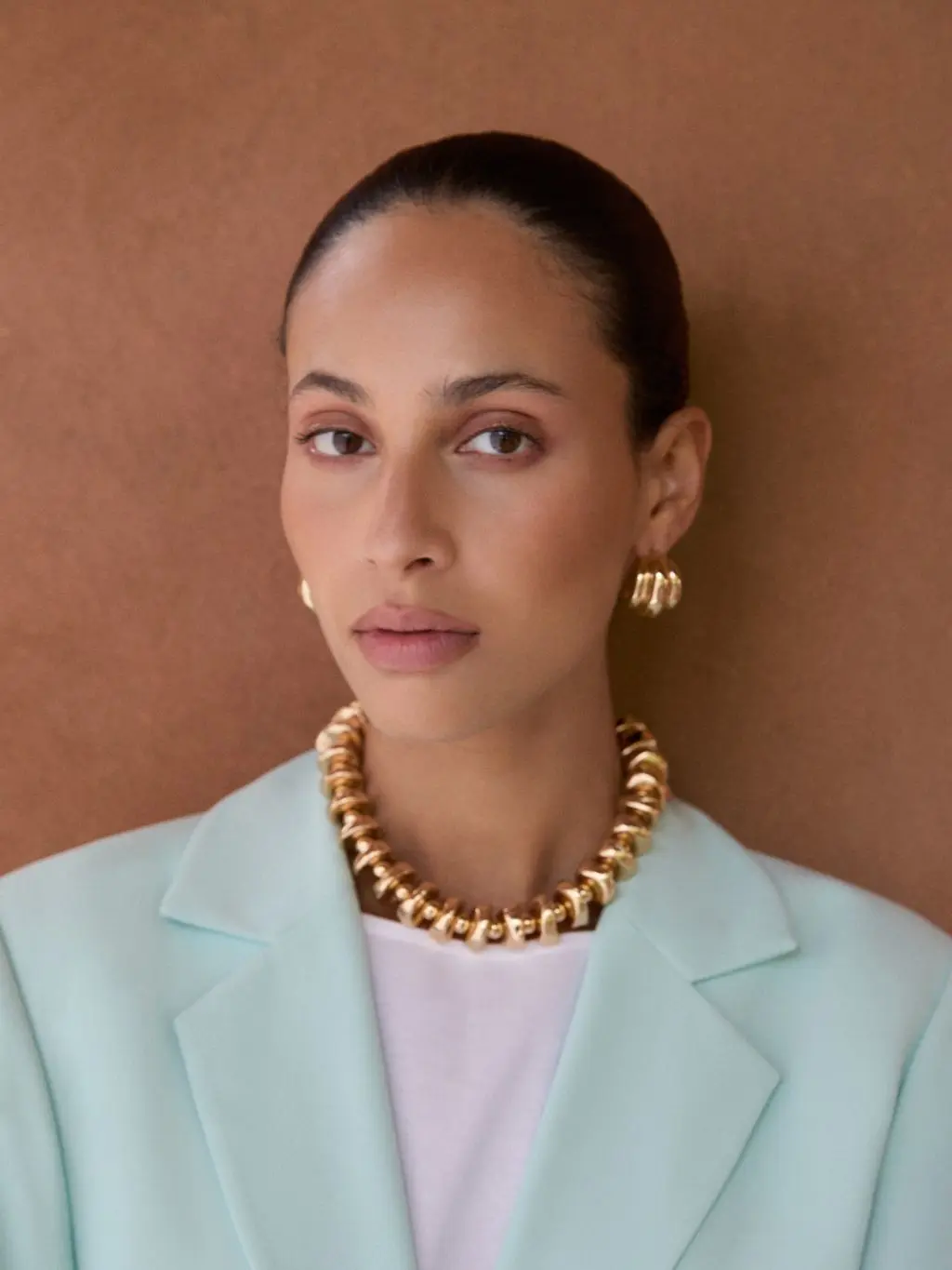 A close up shot of a woman with slicked back hair wearing gold earrings and a gold necklace and a pale blue suit