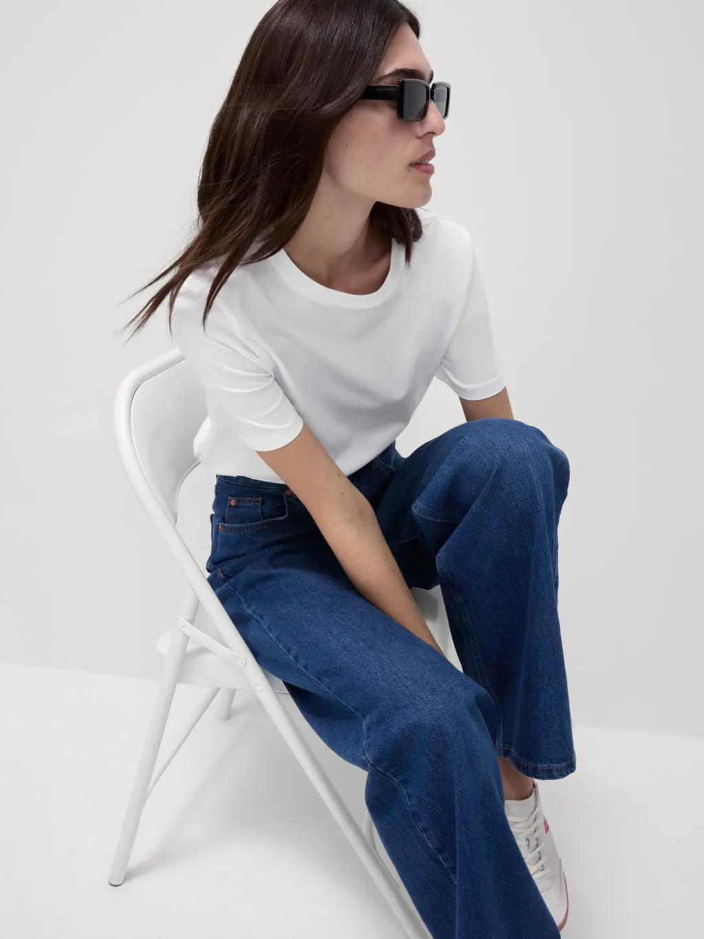 Woman wearing a white T-shirt, dark wash jeans and sunglasses sat on a chair