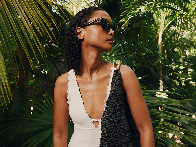 Woman wearing black sunglasses, white scalloped swimsuit and a black beach bag