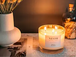 A Neom candle lit and placed on a shelf with a book, matches and dried flowers
