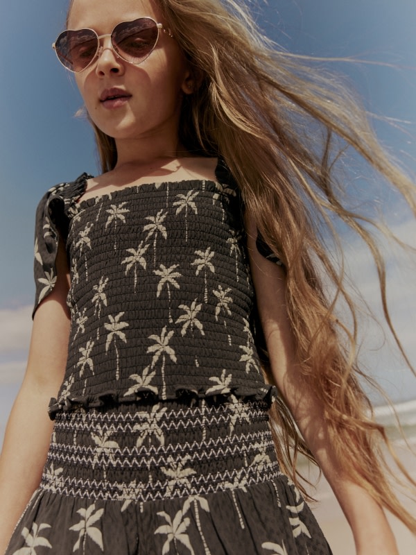 Girl wearing a black and white palm tree printed short-sleeved top and matching skirt. Shop girls’ tops
